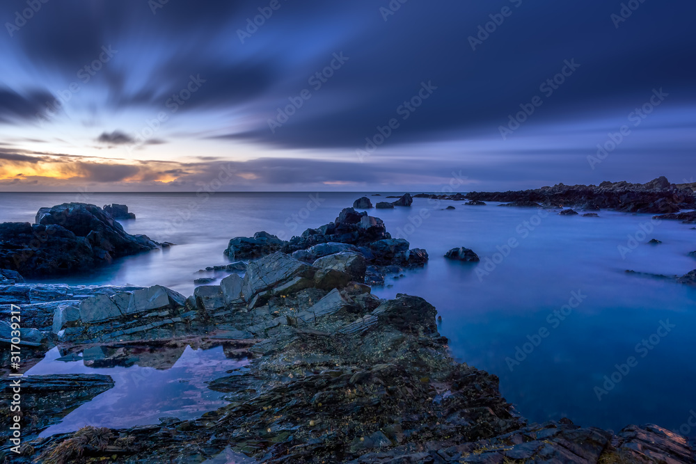 Obraz premium Twilight begins yielding to daylight at rocky coastline with blurred water and sky, long exposure photography. Northern Ireland