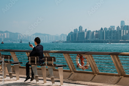 Businessman looking at smart mobile phone outdoors with skyline background, Hong Kong - © hanohiki