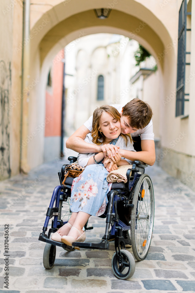 Couple in love in old city center. Pretty young girl with disease on a wheelchair and her lovely man, hugging her from the back Couple in love in old city center. Pretty young girl with disease on a wheelchair and her lovely man, hugging her from the back