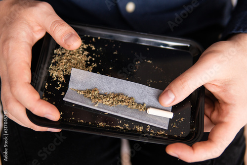 Preparing a joint and drug paraphernalia concept theme with close up man hands rolling a joint with herb girder to grind a cannabis buds in the background