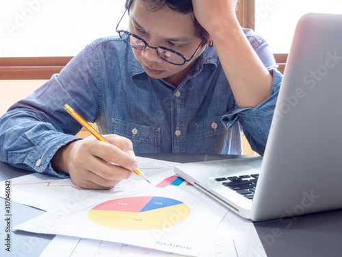 Asian businessman wearing blue shirt and glasses thinking with working with laptop computer on table near the window in office. photo