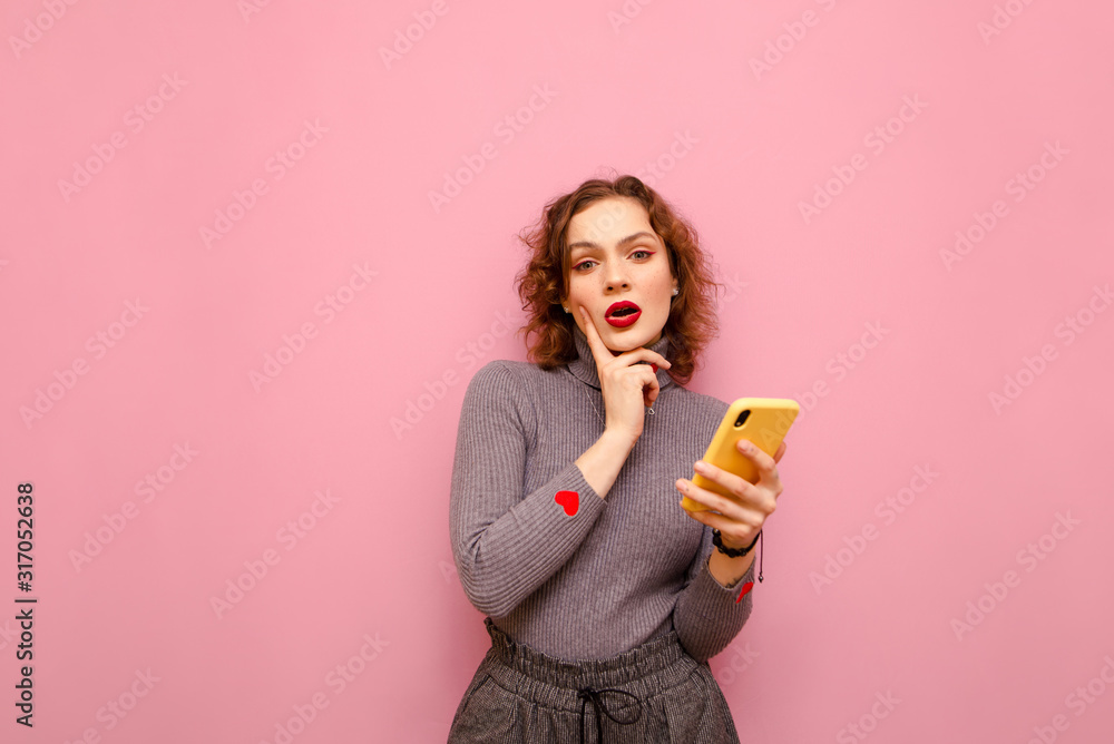 Portrait of pensive lady in casual clothes on pink background, isolated with smartphone in hand, looks into camera and thinks. Smart curly girl in stylish clothes looks at the camera thoughtfully. Portrait of pensive lady in casual clothes on pink background, isolated with smartphone in hand, looks into camera and thinks. Smart curly girl in stylish clothes looks at the camera thoughtfully.