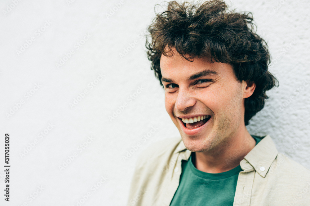 Fototapeta premium Close-up portrait of happy handsome young man smiling broadly posing for advertisement, smiling and looking at the camera. Portrait of attractive male with curly hair has joyful expression.