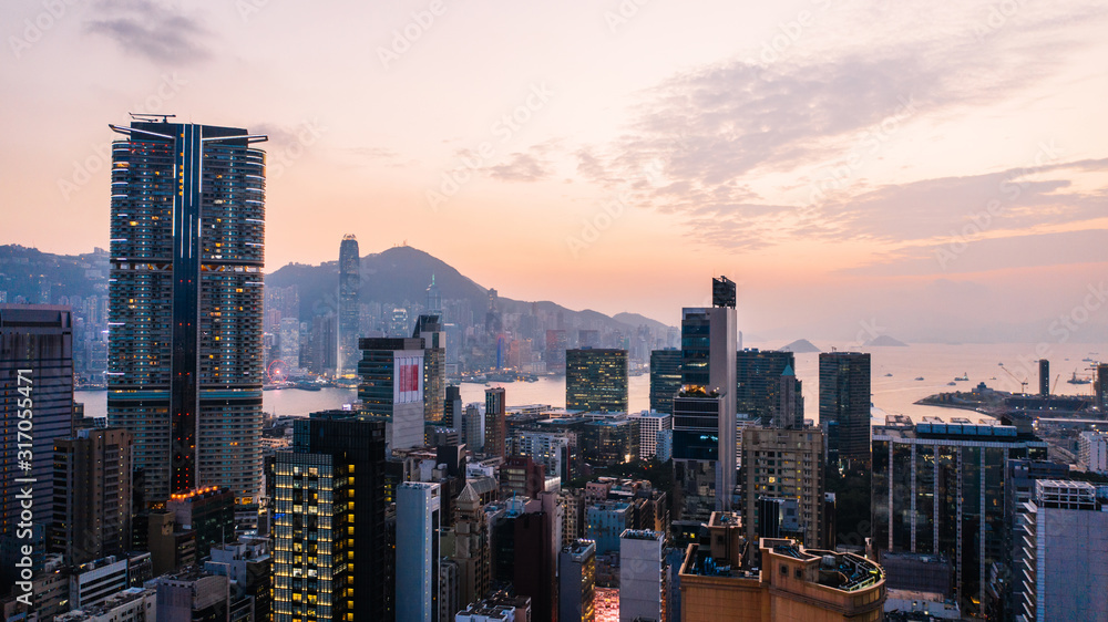 Fototapeta premium Antenowe krajobrazy panoramiczny widok na wieczór w Hongkongu z metropolitalną zatoką Victoria Harbour o zachodzie słońca. Oświetlony nowoczesny pejzaż miejski, budynki miejskie. Infrastruktura energetyczna. Popularne azjatyckie miasto