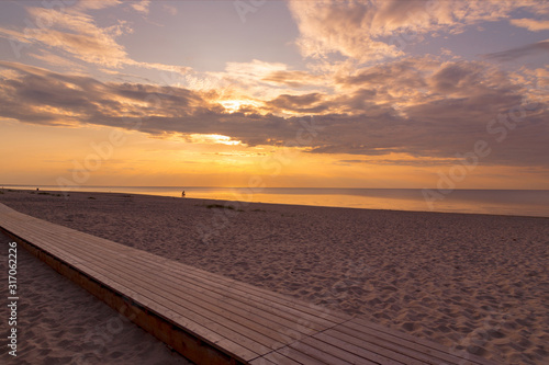 Beautiful sunset sky with clouds over Baltic sea beach coastline. Jurmala. Latvia. Small silhouette of man on a bicycle cycling along a coast. Wooden jetty on foreground.