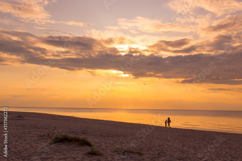 Beautiful sunset sky with clouds over Baltic sea beach coastline. Jurmala. Latvia. Small silhouettes of mother and little son walking along coast.