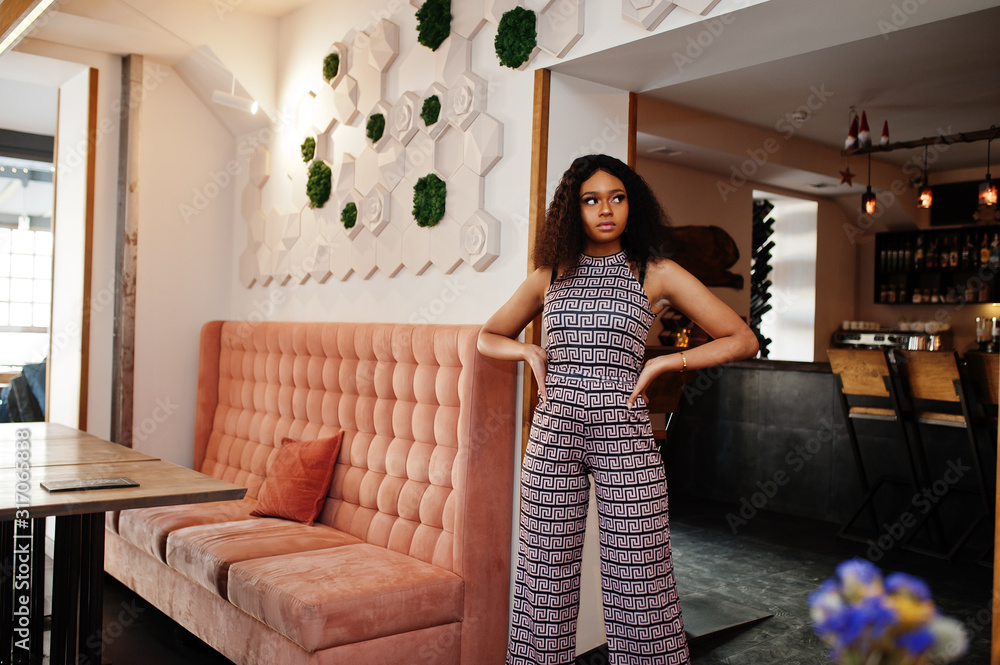 Fototapeta premium Сharming elegant young african american woman with long curly hair wearing jumpsuit posing at cafe indoor.
