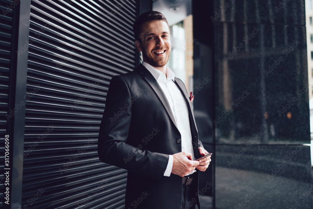Fototapeta premium Pleasant man in classical suit with smartphone looking at camera with smile in street in downtown