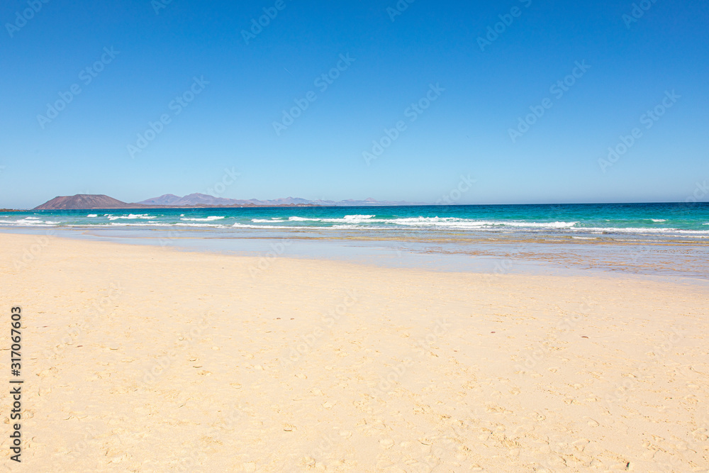 Beach on Fuerteventura canary islands with the beach in front and the sea with blue azure aquamarine turquoise colors and a vulcano in the background blue sky Beach on Fuerteventura canary islands with the beach in front and the sea with blue azure aquamarine turquoise colors and a vulcano in the background blue sky