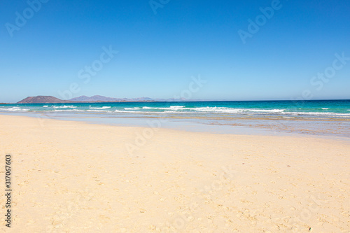 Beach on Fuerteventura canary islands with the beach in front and the sea with blue azure aquamarine turquoise colors and a vulcano in the background blue sky