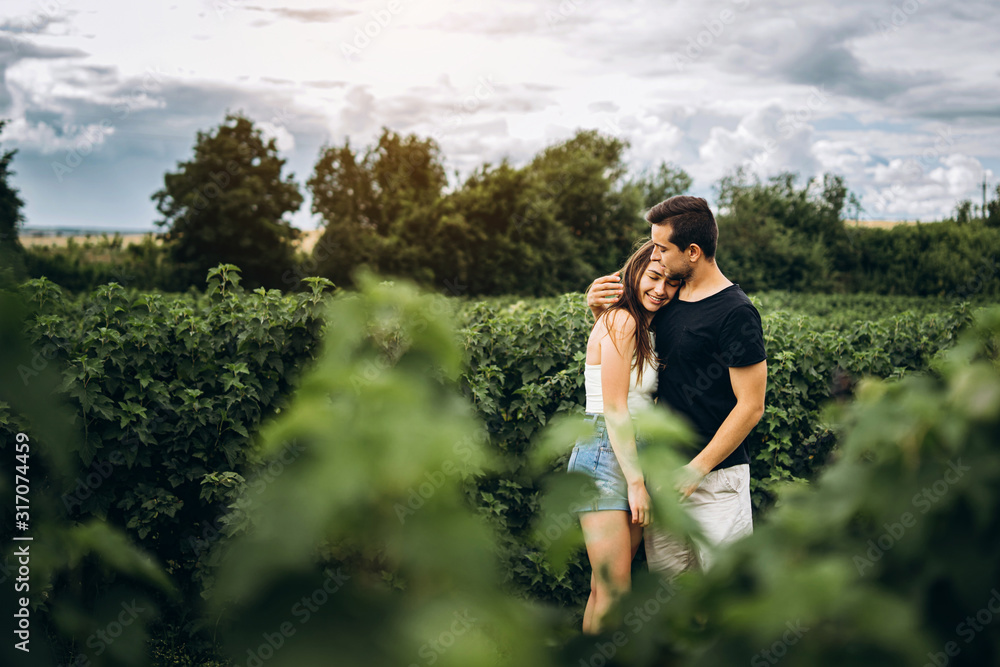 Young loving couple, woman and man, gently hugging with eyes closed on the background of green currant field. Foreground with blurred leaves Young loving couple, woman and man, gently hugging with eyes closed on the background of green currant field. Foreground with blurred leaves