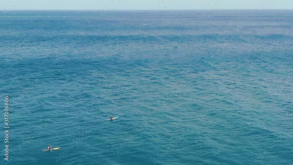 Surfers waiting on their board for big wave to ride, paddling slowly on deep blue ocean near shore of Hawaiian island Surfers waiting on their board for big wave to ride, paddling slowly on deep blue ocean near shore of Hawaiian island