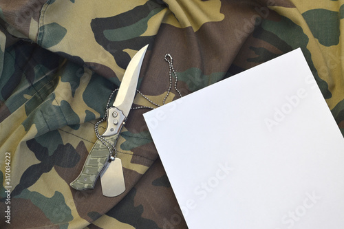 Blank paper with knife and army dog tag lies on camouflage military uniform
