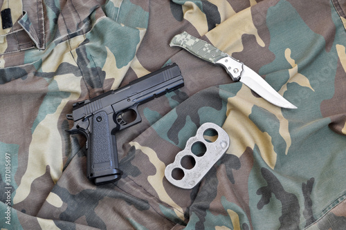 Handgun lies with brass knuckles and knife on camouflage military uniform close up. Concept of looting and arms trade photo