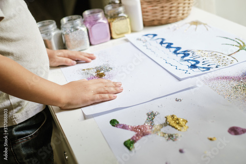 Child's hands and artworks made by child with glue and colourful glitter