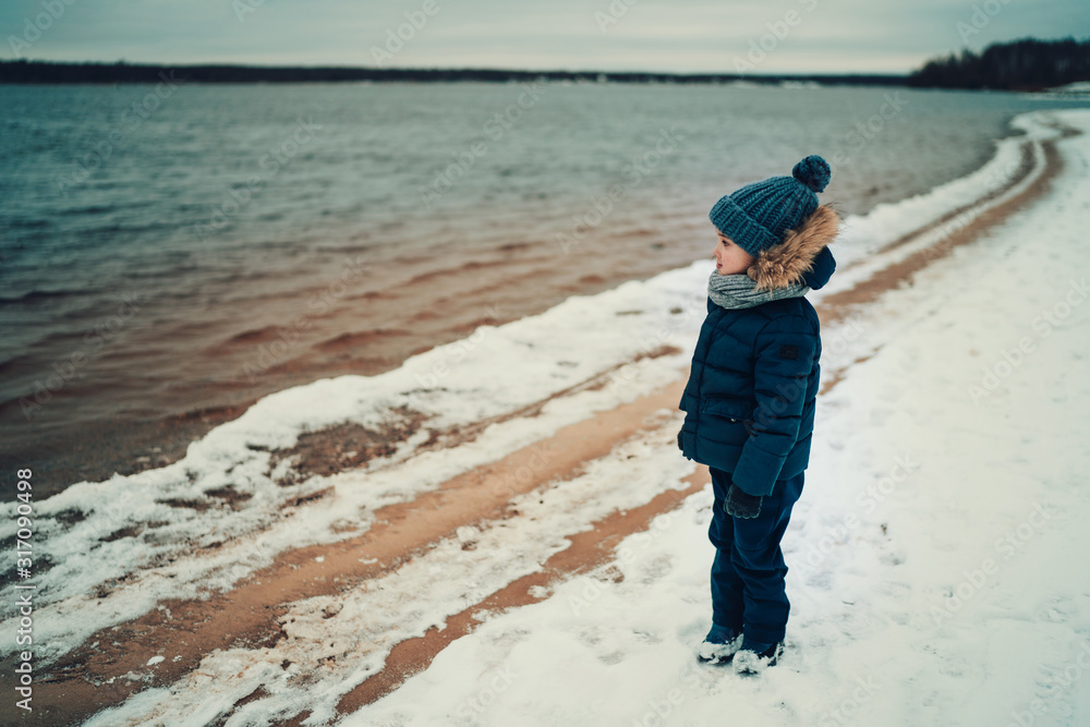 full length shot of small boy looking at the water in the lake on the beach in winter time