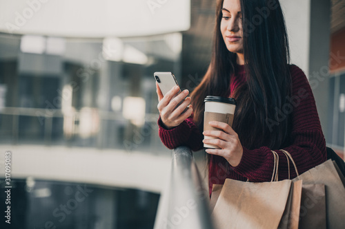 Young pretty woman blogger with smartphone, cup of coffee and shopping bags in city mall