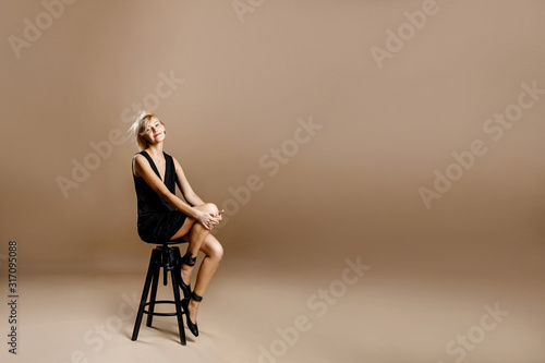 Beautiful blonde woman in a black dress aged 30-40 years old sitting on a chair on a brown background.