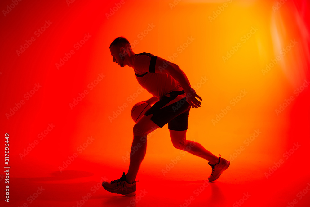 Young athletic man, basketball player dribbling with ball on red and orange background