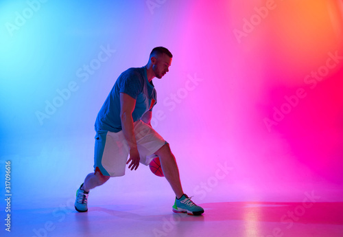 Young athletic man, basketball player dribbling with ball on neon blue and pink background
