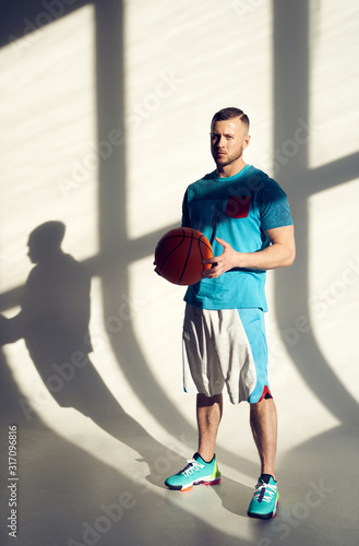 Young athletic man, basketball player holding ball and standing near wall with shadows from window
