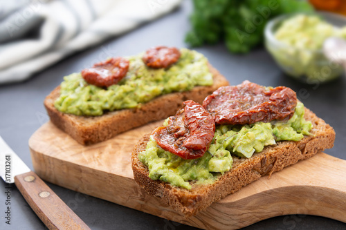 Two avocado sandwiches. Rye bread with guakomole, avocado pasta and dried tomatoes, on wooden cutting board. Avocado toast.