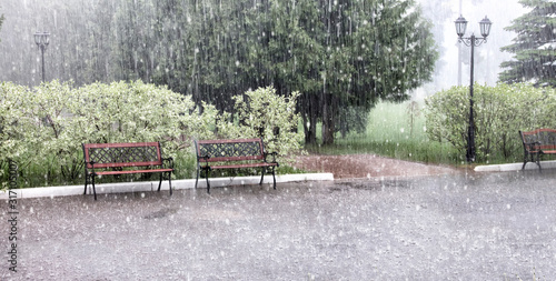 Rain and hail on a summer day. Large puddles on the sidewalk during heavy rain. Heavy rain with snow and hail in the Park. Wet benches on the paved path. Spray from drops and bubbles in large puddles.
