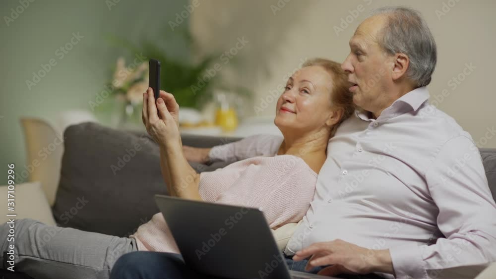Loving senior couple relaxing at home together. Man sitting on sofa and using laptop computer, wife leaning on him and showing pictures on smartphone Loving senior couple relaxing at home together. Man sitting on sofa and using laptop computer, wife leaning on him and showing pictures on smartphone