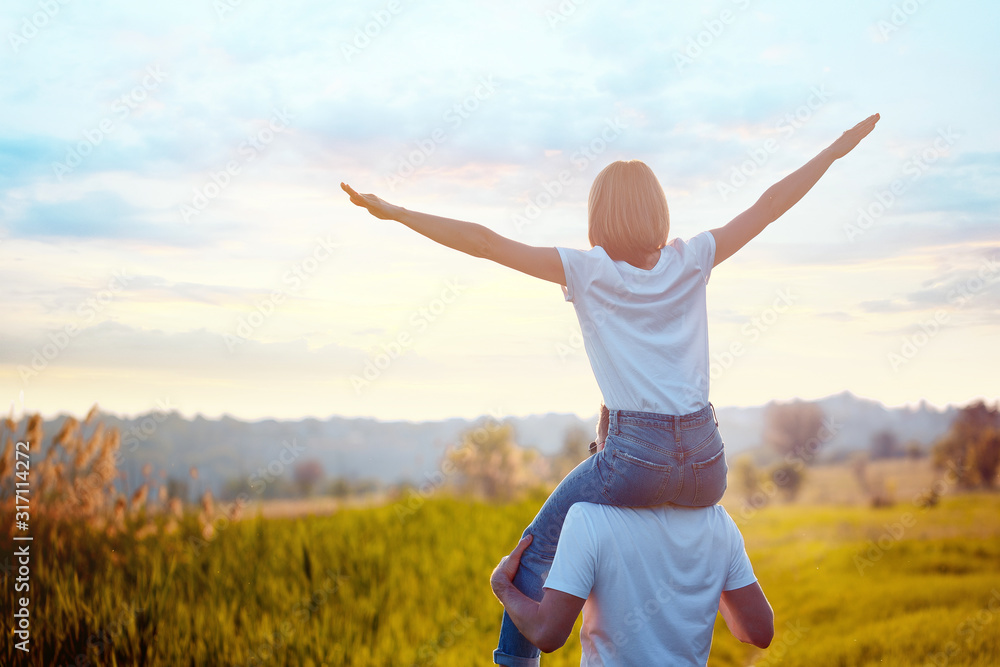 Fototapeta premium Portrait of young man holding on shoulders his happy girlfriend, she spread apart arms mimicking airplane. Summer. Vacation air travel concept. Sunset.