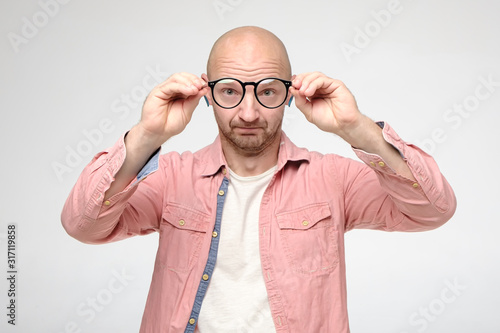 Bald, unshaven man took off glasses, keeping them at a distance, he examines the cleanliness of the lenses and looks funny. © koldunova