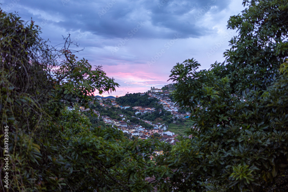 Early morning colonial mining town Ouro Preto in Minas Gerais, Brazil, with clouds reflecting a colourful pink and orange sunrise seen through vegetation in the foreground