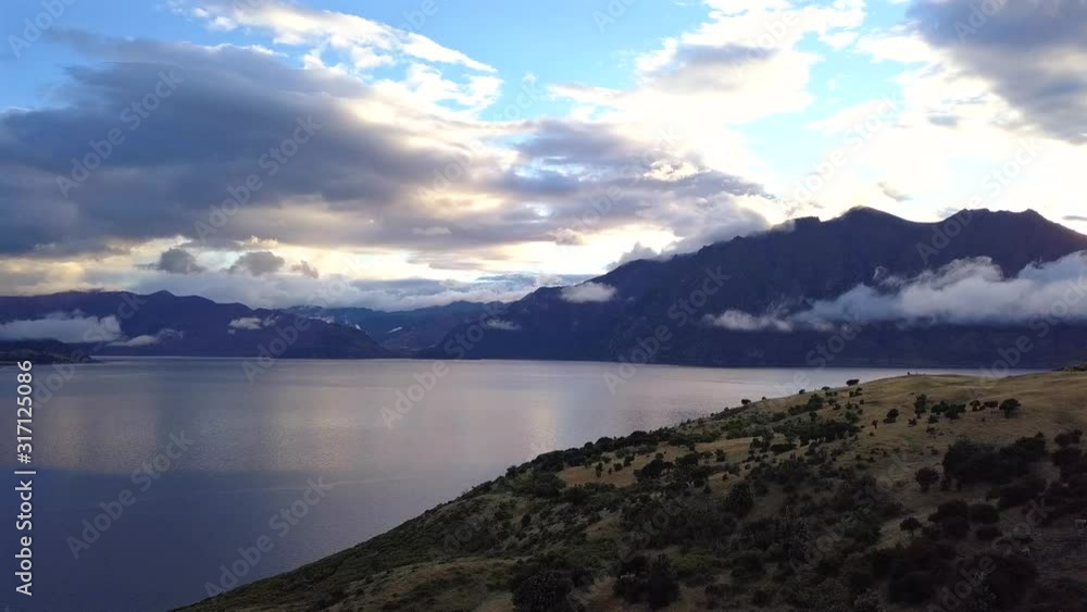 A simple aerial jib shot highlights the scale of a large New Zealand lake and low cloud cover at sunrise, against a foreground of sparsely foliaged hillside A simple aerial jib shot highlights the scale of a large New Zealand lake and low cloud cover at sunrise, against a foreground of sparsely foliaged hillside