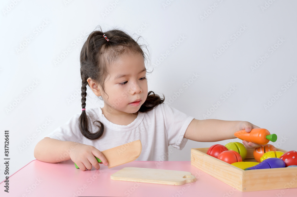 Cute asian girl play cooking at home. Children are using wooden toy knife slicing vegetable on chopping wood board. Education and learning Concept. Cute asian girl play cooking at home. Children are using wooden toy knife slicing vegetable on chopping wood board. Education and learning Concept.