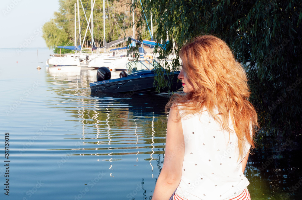 Fototapeta premium back view of beautiful woman with long hair in white blouse sitting near river water with boats and looking away in yachts club