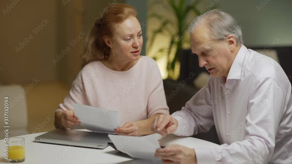 Puzzled elderly couple sitting at table at home and calculating home finances together. Wife and husband holding paper bills and counting expenses on calculator Puzzled elderly couple sitting at table at home and calculating home finances together. Wife and husband holding paper bills and counting expenses on calculator