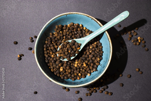 Black peppercorns in blue bowl on dark background, flat lay pepper grains, spoon in bowl