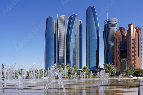view of a row of scyscraper in abu dhabi behind a fountaion, beautiful blue sky and green trees, with some reflections in the water, skyline