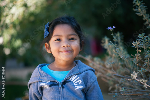 Confident little girl wearing a sweater, standing in front of trees and green plants with a sincere smile on her face.