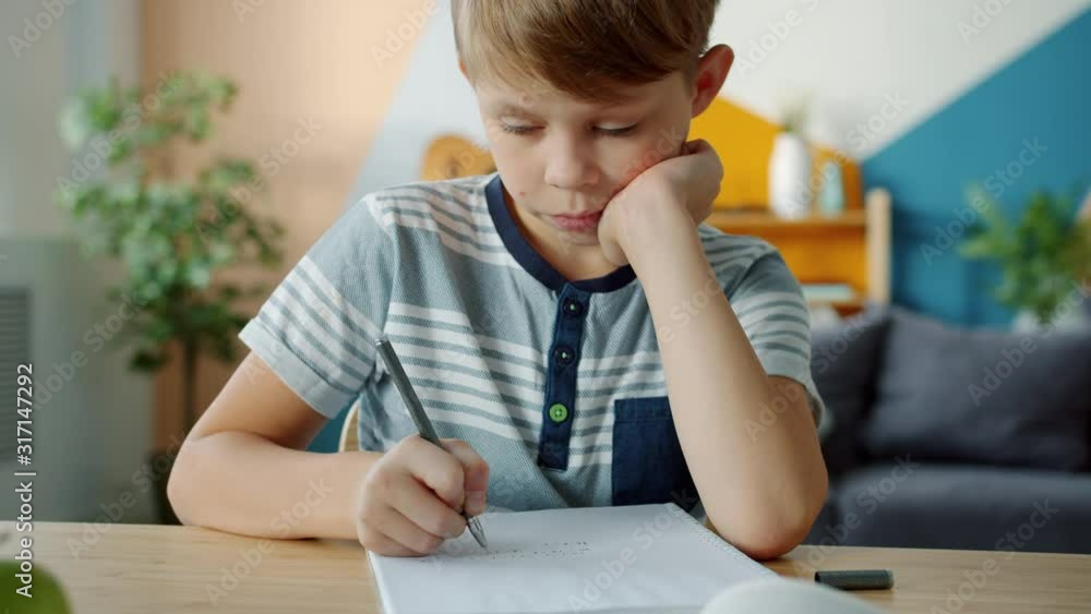 Serious child is doing homework writing in notebook sitting at desk in apartment learning busy with self-education. People, childhood and lifestyle concept. Serious child is doing homework writing in notebook sitting at desk in apartment learning busy with self-education. People, childhood and lifestyle concept.