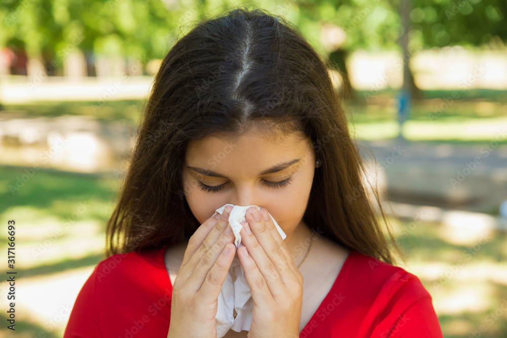 Unhealthy brunette girl blowing nose into tissue. Pretty female having flu or allergy against blurred green spring park background. Sickness, allergy concept Unhealthy brunette girl blowing nose into tissue. Pretty female having flu or allergy against blurred green spring park background. Sickness, allergy concept