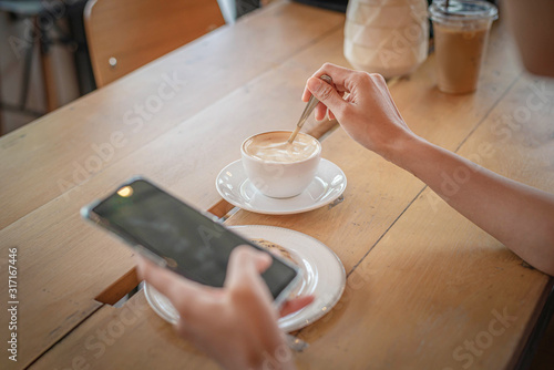 A girl sitting in a coffee shop at a wooden table drinking coffee and using a smartphone on the table as a laptop Girl surfing the internet chat blog With friends on the phone and watch on his screen © Tum