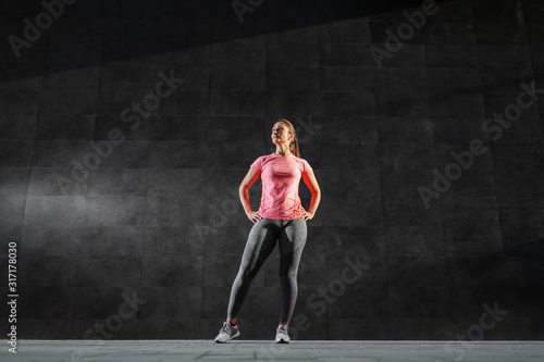 Full length of fit attractive caucasian brunette in sportswear and with ponytail standing with hands on hips and looking away. In background is dark wall.