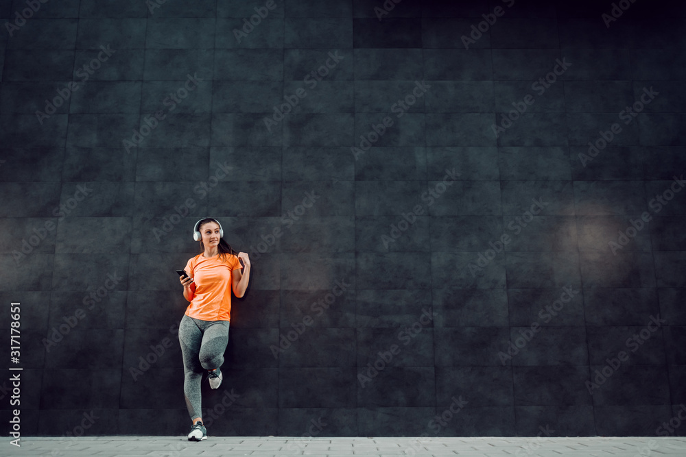 Φωτογραφία Full length of attractive caucasian sportswoman in shape leaning on dark wall, holding smart smart phone, listening music and looking away
