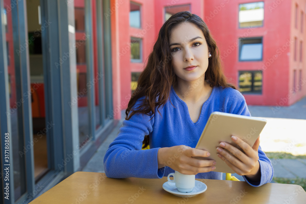 Calm young woman holding tablet and looking at camera. Front view of tranquil lady sitting at table at outdoor cafe. Technology concept Calm young woman holding tablet and looking at camera. Front view of tranquil lady sitting at table at outdoor cafe. Technology concept