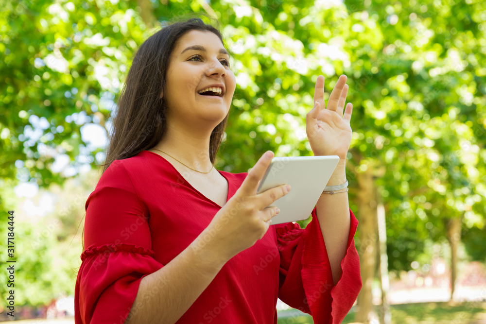 Smiling teen girl holding tablet and waving. Cheerful teenager standing in spring park and holding modern tablet. Technology concept Smiling teen girl holding tablet and waving. Cheerful teenager standing in spring park and holding modern tablet. Technology concept