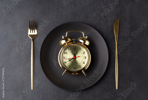 Golden alarm clock on a black plate with a golden knife and fork