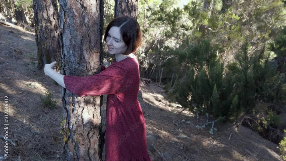 Pretty happy adult caucasian environmental woman in red dress hugging tree in forest during sunset Pretty happy adult caucasian environmental woman in red dress hugging tree in forest during sunset
