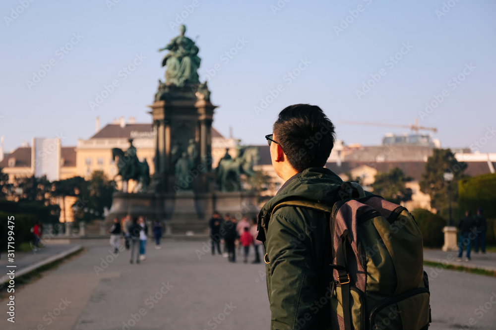 Fototapeta premium Back view portrait of Asian male tourist backpacker carrying a bag in Maria Theresa Monument in Vienna, Austria, Europe