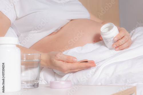 Unrecognizable Pregnant Woman in bed holding vitamins, pills in hand before intake. Glass of water is on bedside table. Pregnancy healthy lifestyle. Snow-white bedding.