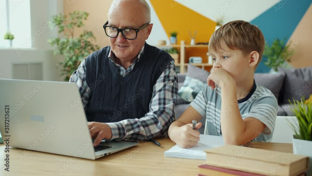 Old man is using laptop computer helping child with homework at desk in apartment, boy is pointing at screen talking. Technology and childhood concept. Old man is using laptop computer helping child with homework at desk in apartment, boy is pointing at screen talking. Technology and childhood concept.